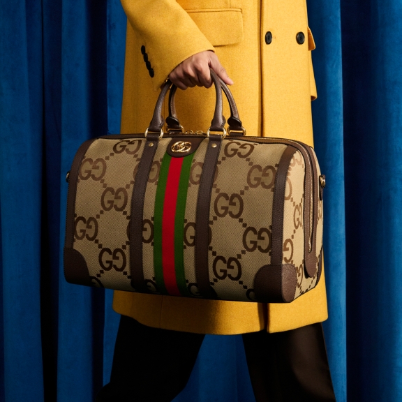 The image features a person holding a luxury Gucci bag, prominently displaying a distinctive logo pattern with green and red stripes. The person is also wearing a chic mustard yellow coat against a blue backdrop.