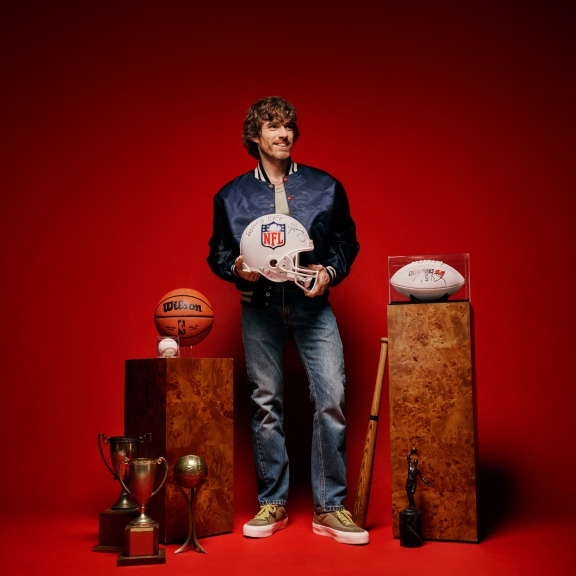 A collector stands among two pedestals with rare sport memorabilia collectibles around them. They stand in front of a red seamless background.