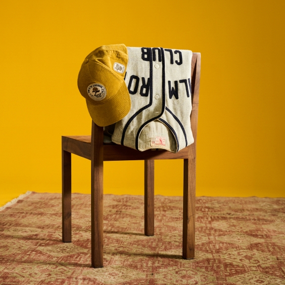 An old baseball jersey and a yellow cap rest on the back of a wooden chair. The wooden chair is on top of a red decorated rug on a yellow seamless background.