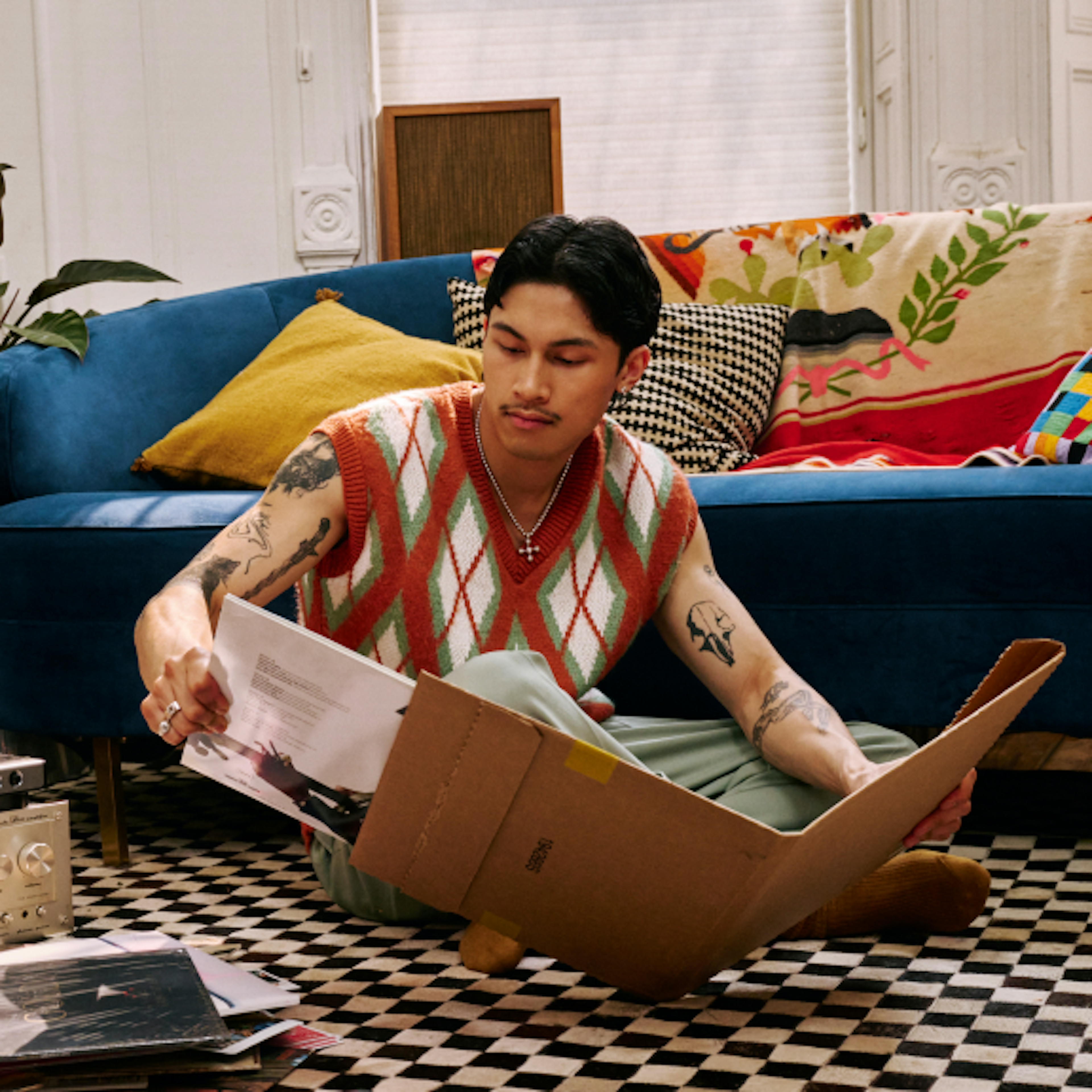 The image features a man sitting on a checkered floor, looking through a box of papers in a cozy, vibrant living room. He wears a stylish, patterned vest and has visible tattoos, adding to his artistic vibe. The room is decorated with colorful cushions and a blue sofa, giving it a warm, eclectic feel.