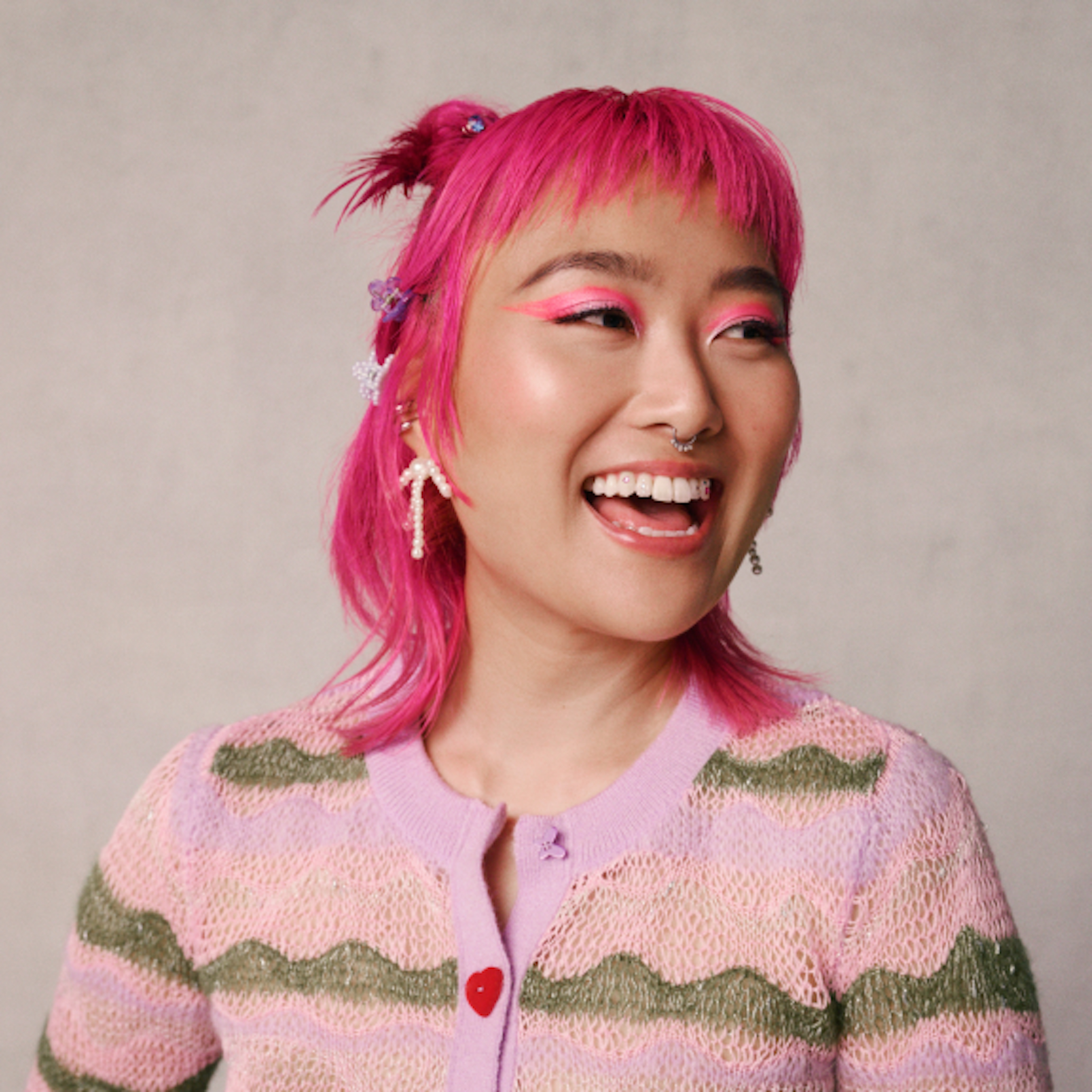 The image shows a woman with a joyful expression, sporting vibrant pink hair styled with small decorations. She wears a delicate pink cardigan with a green wavy pattern. Her makeup complements her hair with bright pink tones, and she has a cheerful, engaging smile. The background is a simple, neutral tone that highlights her colorful appearance.