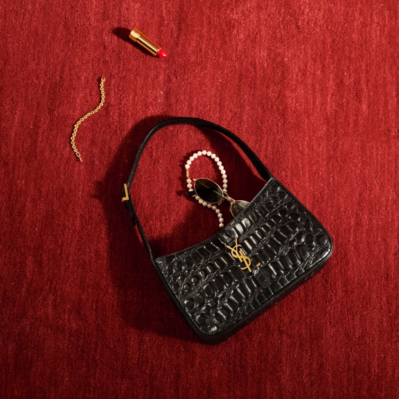 The image shows a YSL purse on a red carpet styled with pearls, sunglasses, and an open lipstick spilling out of it as if recently dropped.