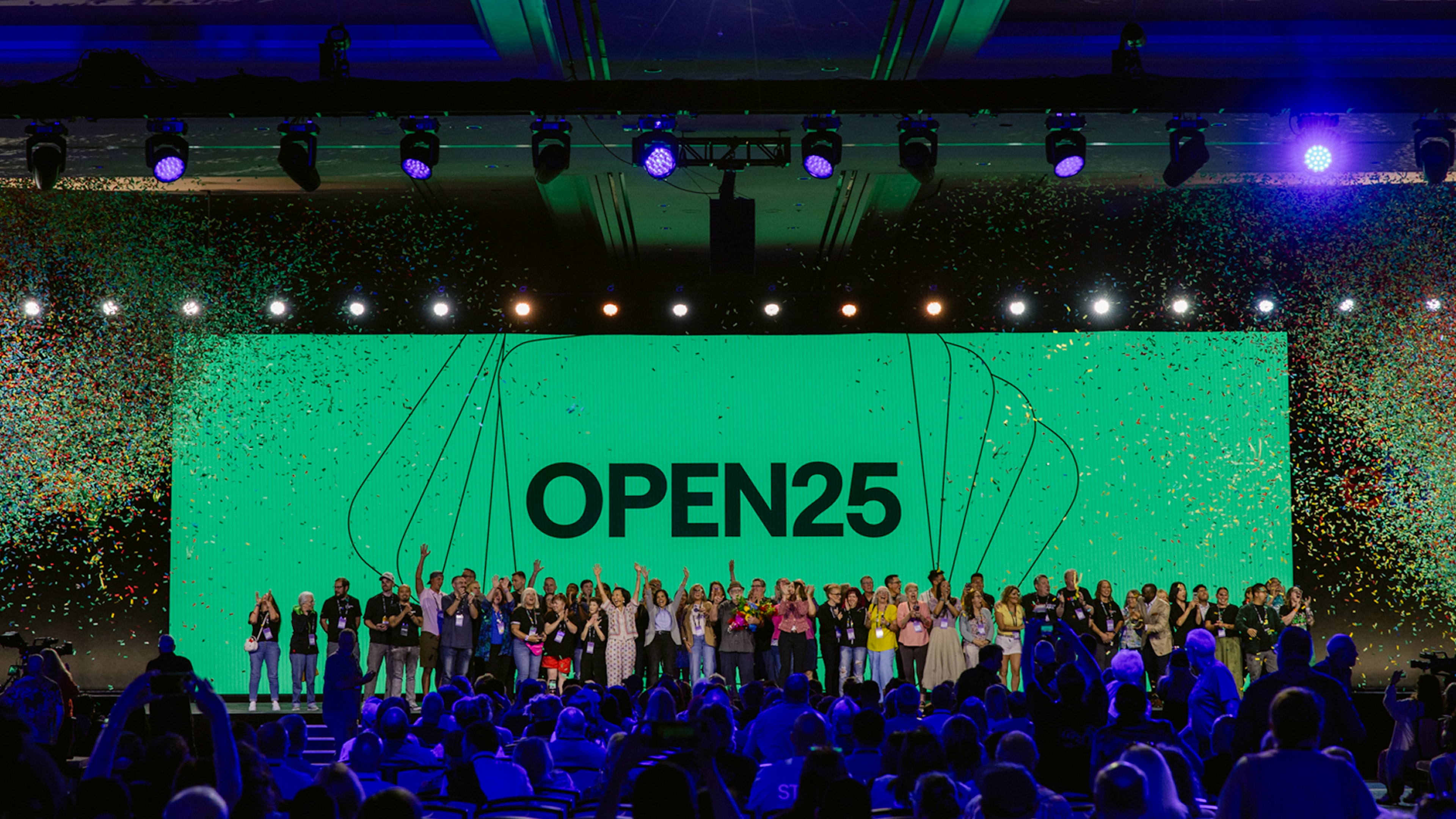 A large group of people on stage celebrating in front of a bright green screen reading “OPEN25,” with confetti falling and an audience watching under colorful stage lights.