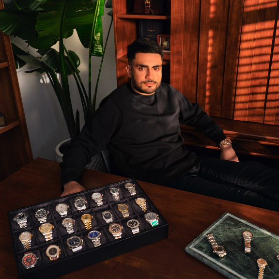 A watch seller sits at a desk in a warmly lit showroom, with display trays of luxury watches arranged in front of him and additional timepieces showcased on shelves behind him.