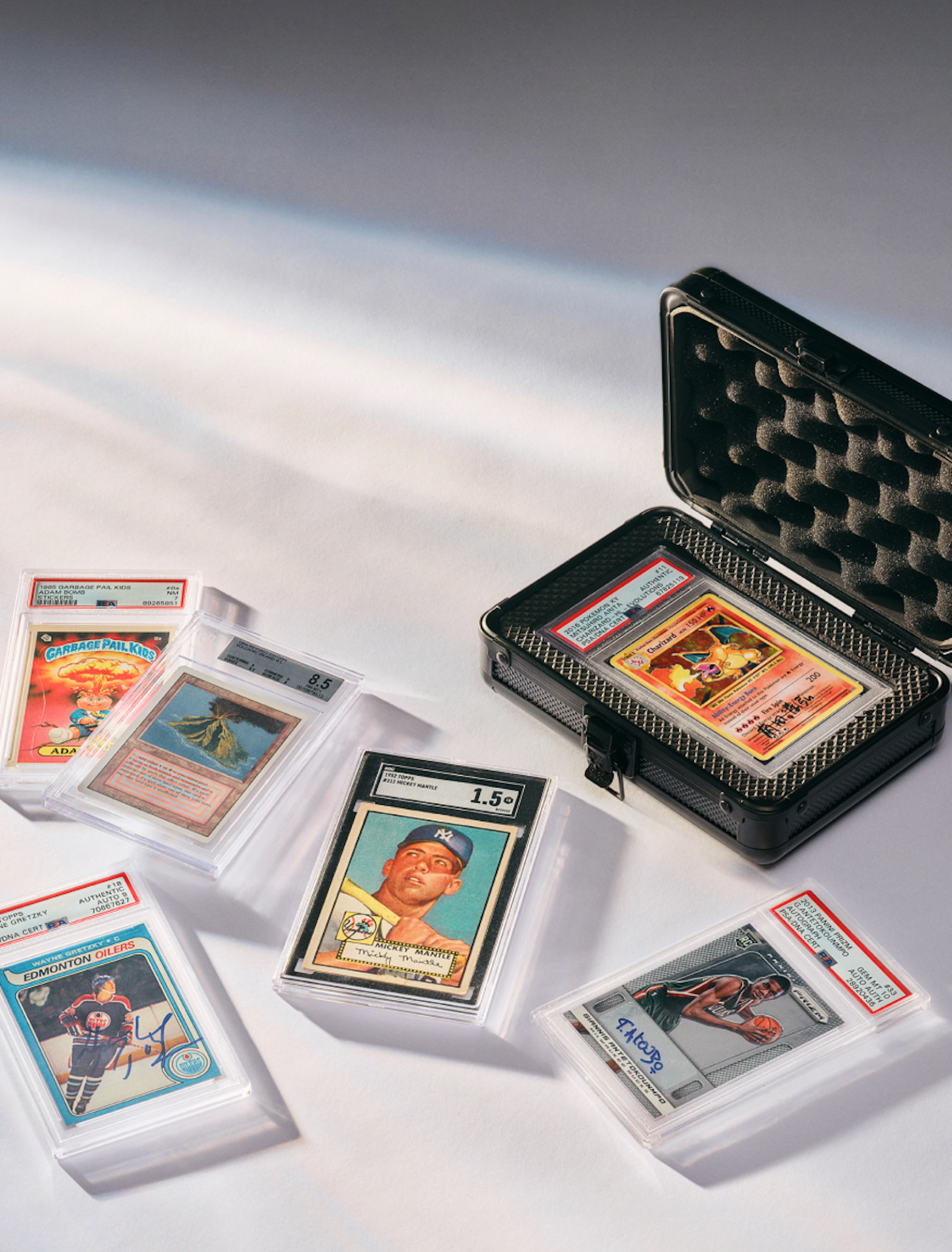 A collection of professionally graded sports and trading cards is displayed on a white surface, with one featured card secured inside an open protective hard case.