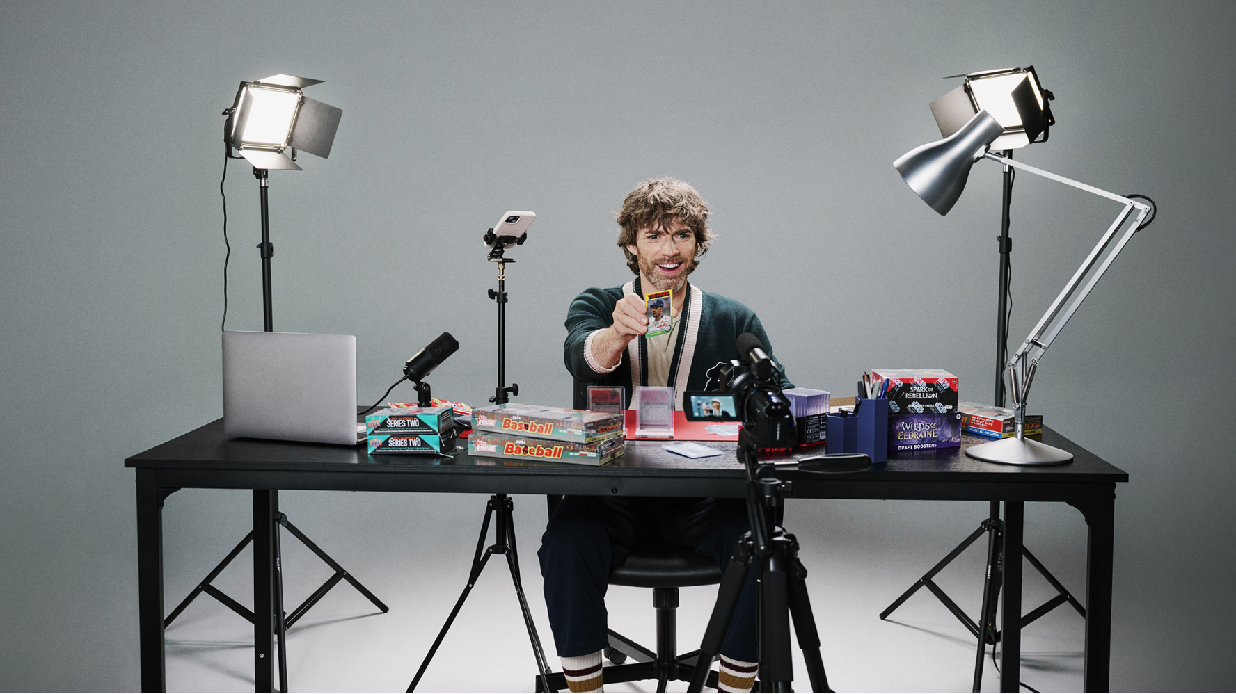 A trading card seller sits at a eBay LIVE studio setup with cameras, lights, and a laptop, holding up a collectible card while livestreaming or recording content at a desk covered with trading card products and supplies.