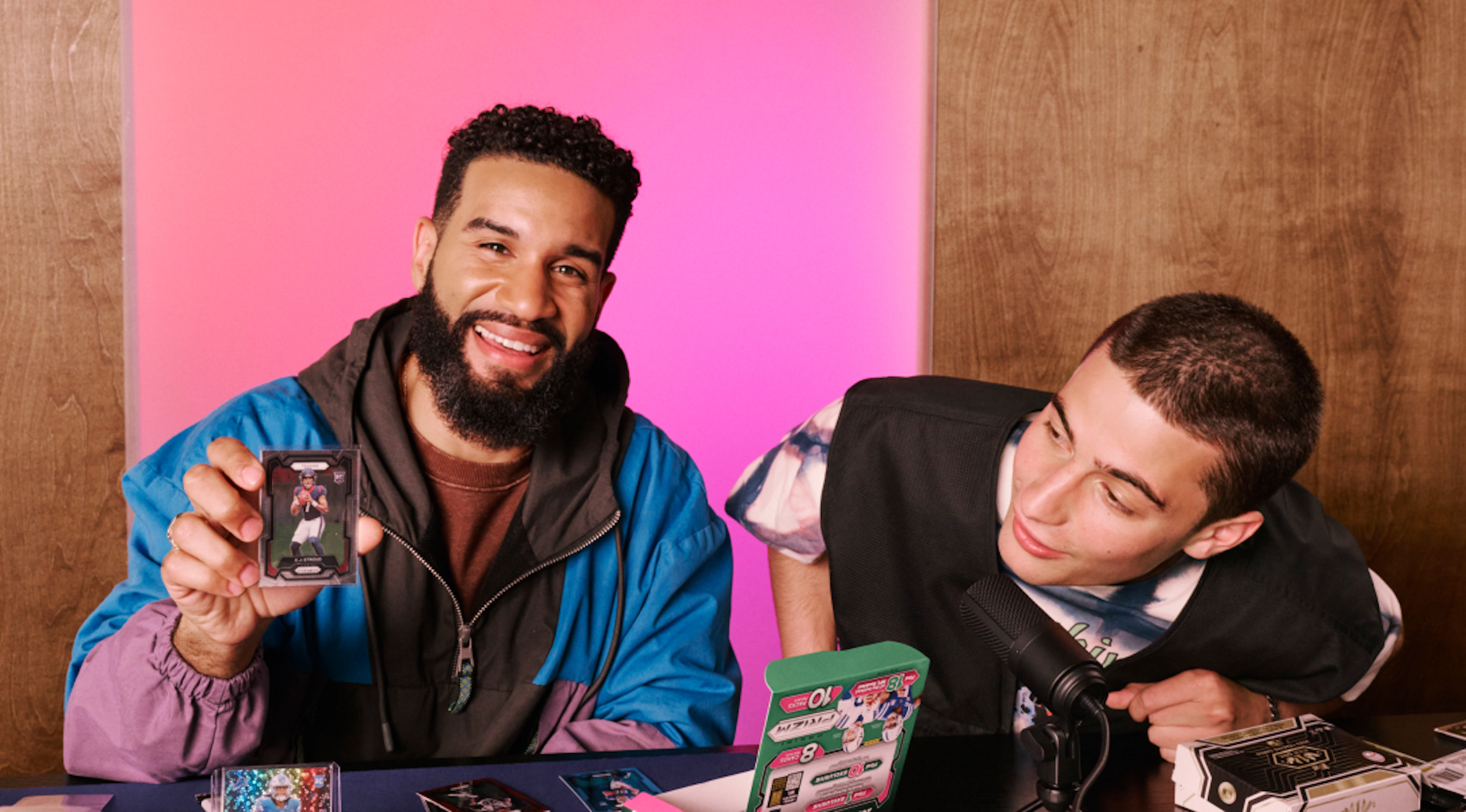 Two trading card collectors sit at a table with a microphone and card packs, smiling as one proudly holds up a collectible card while the other leans in to look.