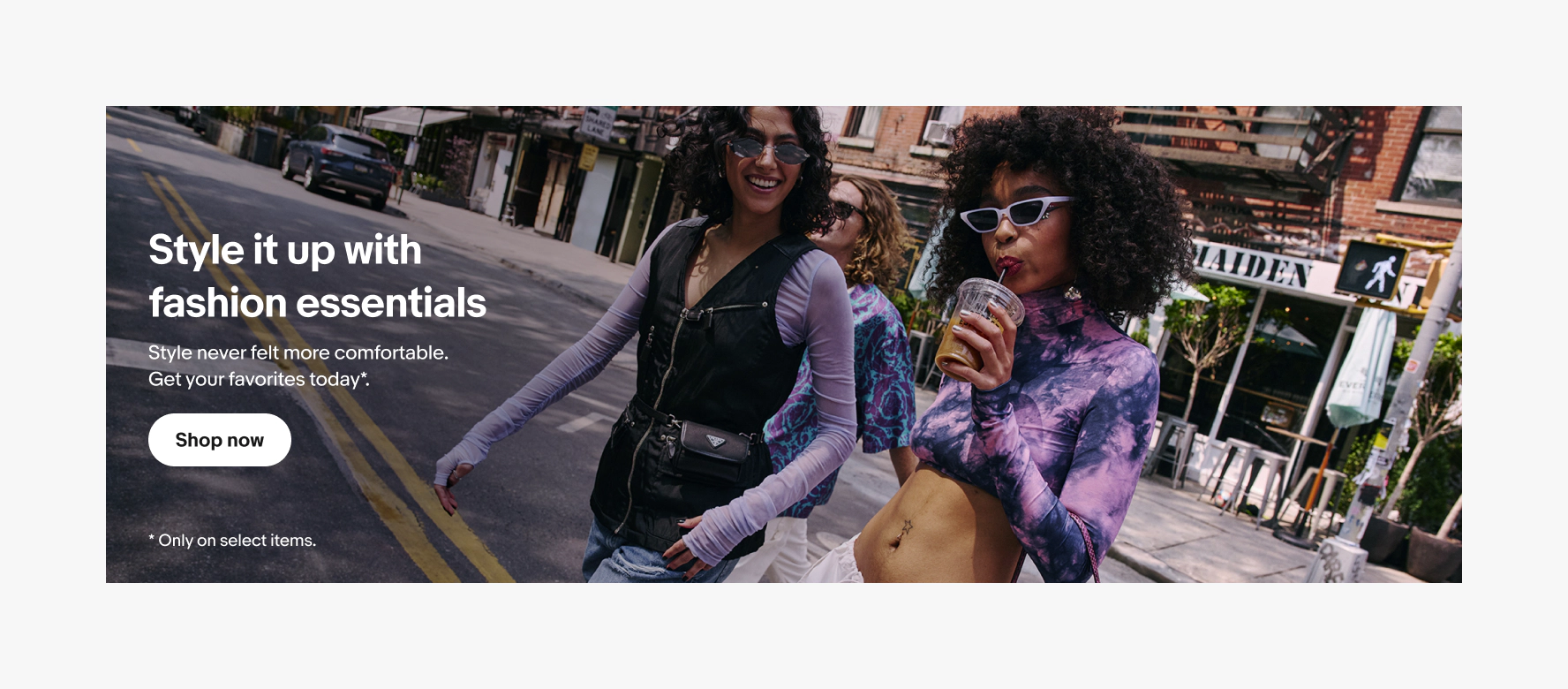 Banner with a full-bleed photo background and white text centered vertically on top. The photo is of three stylish friends walk down a city street wearing colorful layered outfits and sunglasses. Text reads, “Style it up with fashion essentials,” with a “Shop now” button.