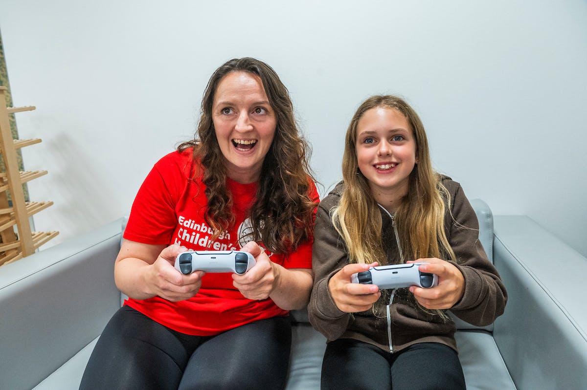 Women in a red Edinburgh Children's Hospital t-shirt and a girl holding a game controller sat on a grey sofa.