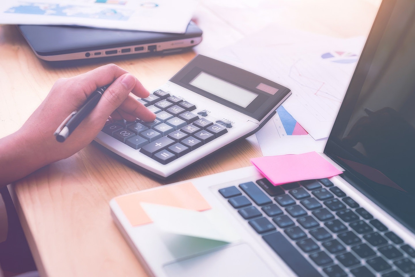 Person using a calculator and pen at a desk with a laptop and printed charts, suggesting financial or operational analysis.