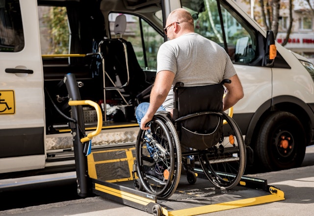 Wheelchair user positioned on a lowered platform lift entering an accessible transit van parked at the curb.