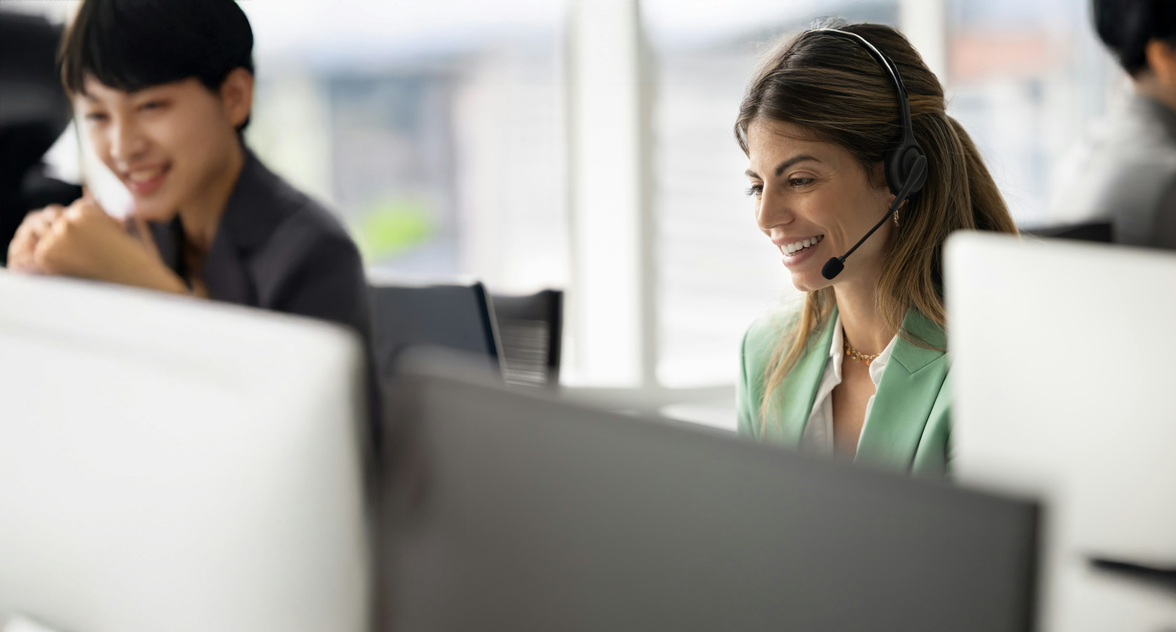 Customer support agents wearing headsets work at their desks in a bright office, assisting customers with friendly, attentive expressions.