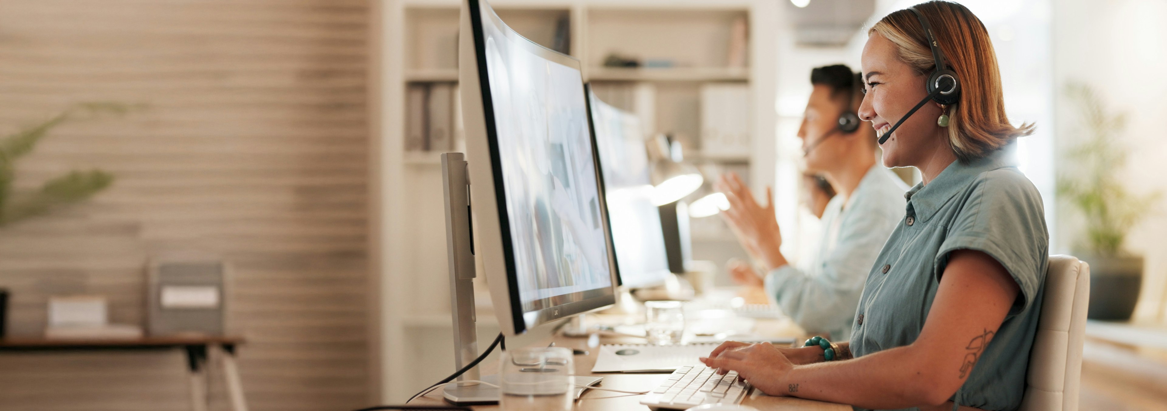 Customer support agents wearing headsets work at desktop computers in a modern office, assisting customers through live calls.