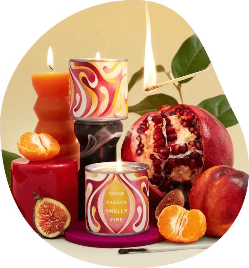 A photograph showing the Daye Vagina Smells candle alongside other red candles a tangerine a fig a pomegranate and a peach as well as a green plan in the background and a lit match