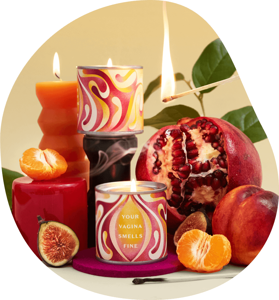 A photograph showing the Daye Vagina Smells candle alongside other red candles a tangerine a fig a pomegranate and a peach as well as a green plan in the background and a lit match