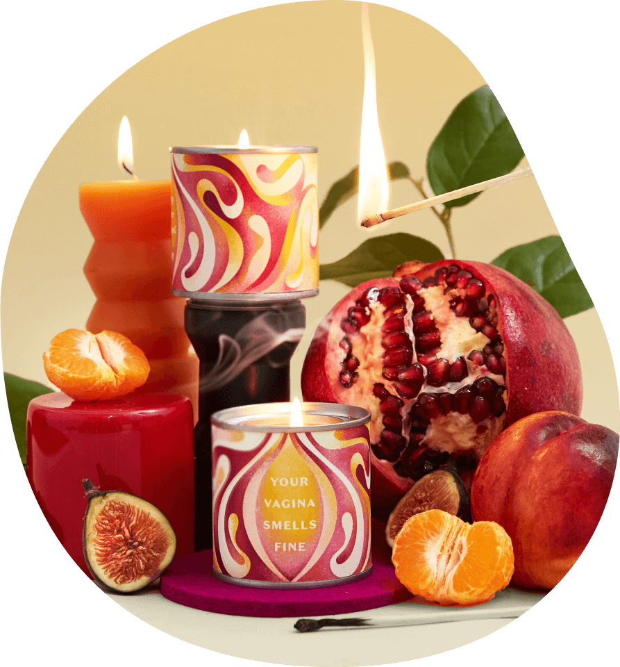 A photograph showing the Daye Vagina Smells candle alongside other red candles a tangerine a fig a pomegranate and a peach as well as a green plan in the background and a lit match 