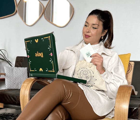 A woman in cream shirt with chocolate leather pants holding a green box of Daye pads sitting on a chair 