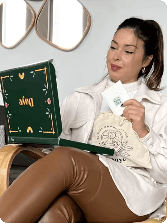 Woman sitting on a chair with a green Daye box on her lap, holding a Daye pad. She is dressed in a light beige jacket and brown leather pants.