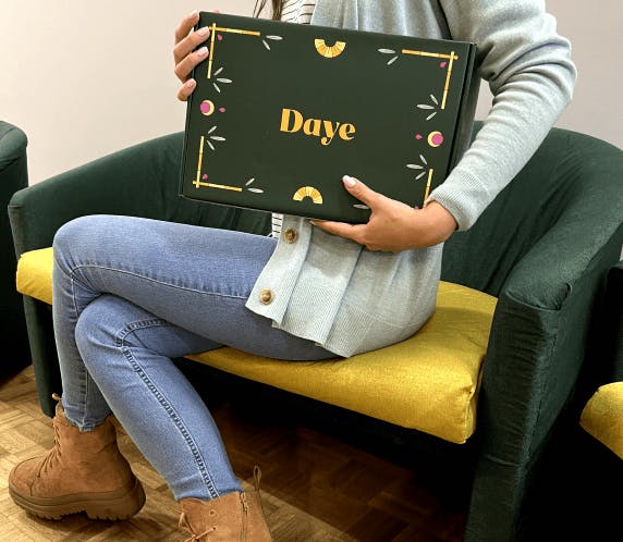 A woman in blue jeans is sitting on a green sofa and is showing a Daye pads box for bamboo liners, super and regular pads