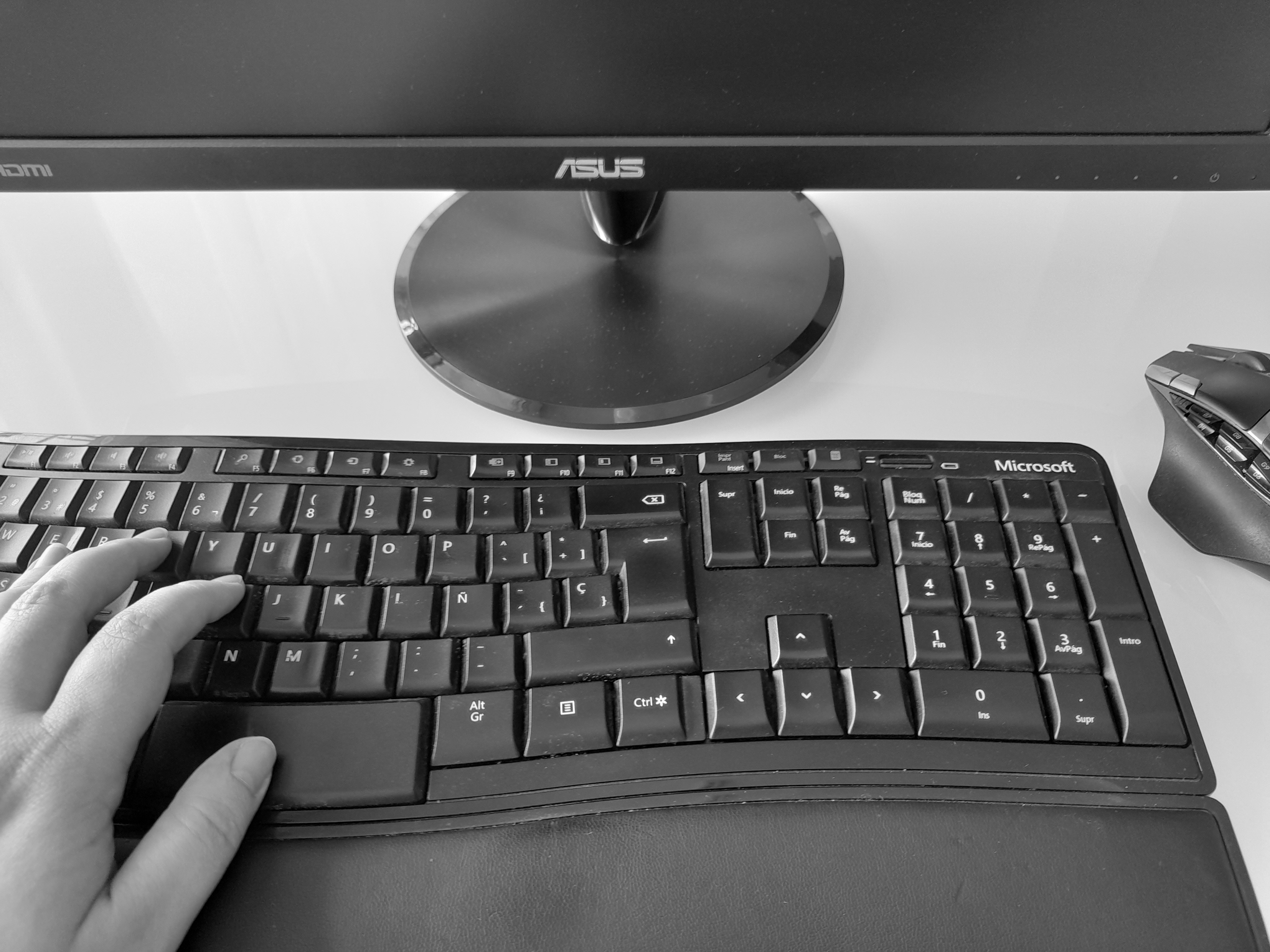 Detal d'un teclat d'ordinador amb una mà que escriu. A la dreta es veu part d'un ratolí, dalt una pantalla.