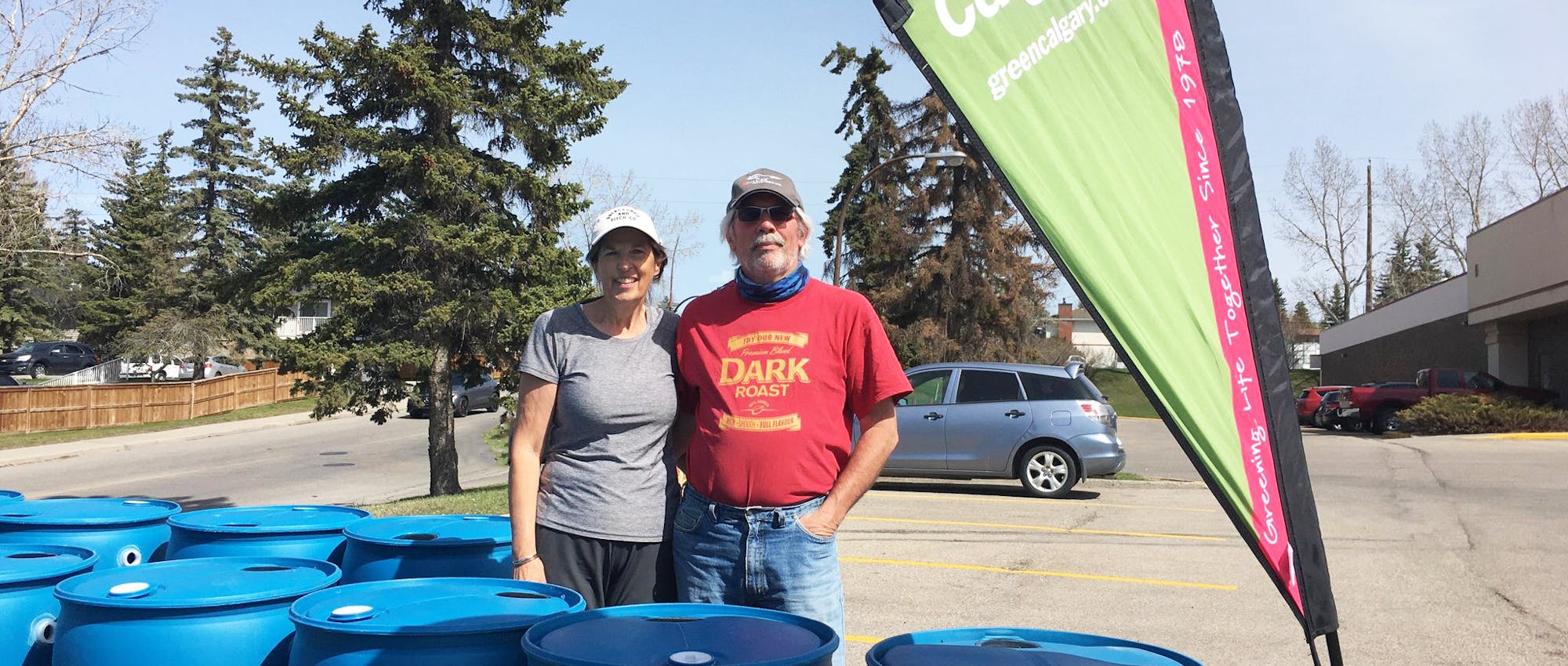 Two people standing beside a Green Calgary flag and rain barrels.