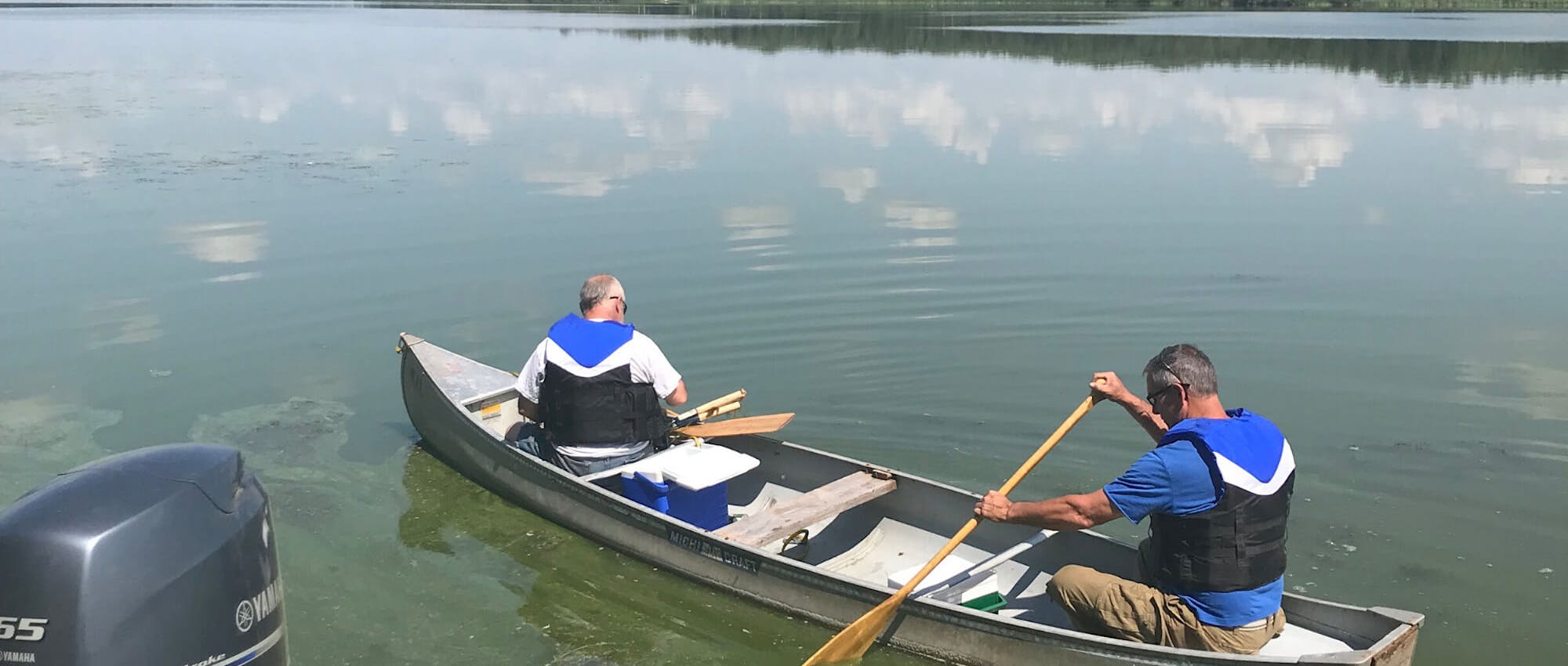 Two men in blue outfits sit in a canoe in a lake.