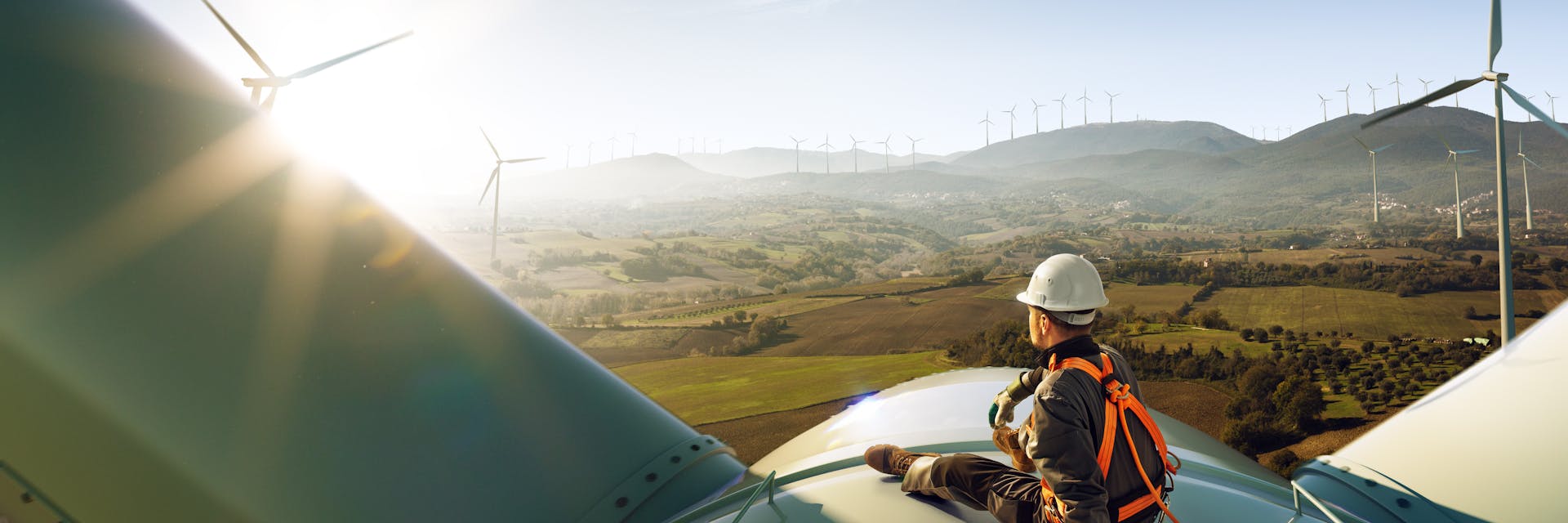 A man in a hard hat sits on top of a windmill in a windfarm.