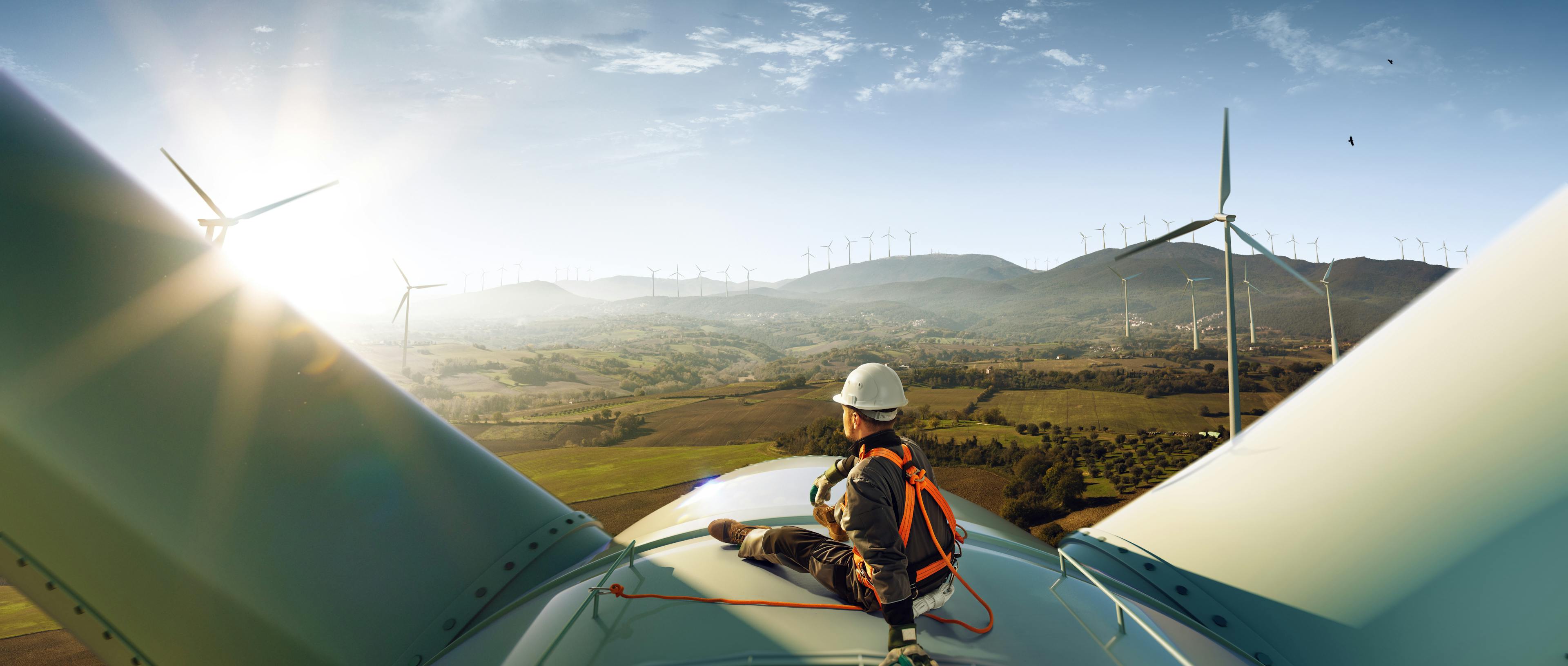 A man in a hard hat sits on top of a windmill in a windfarm.