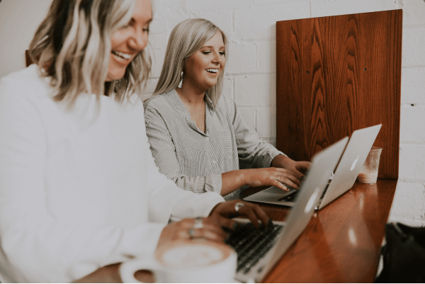 two women working on their laptops side-by-side smiling and laughing