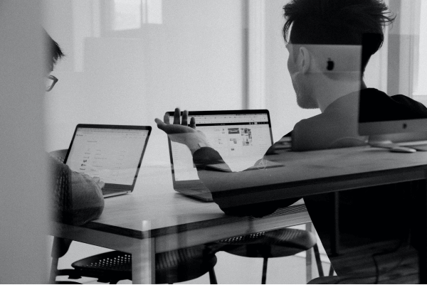 Black and white photo of two people inside a glass meeting room discussing, negotiating