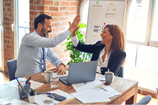 male-coded person high fiving a female-coded person over a meeting table, lots of light, both are smiling