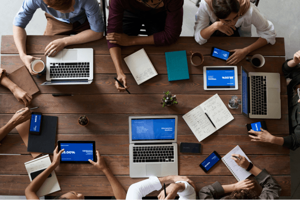 Team huddle together on a wooden table with laptops, tablets, and phones out