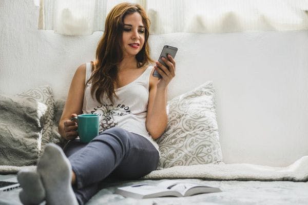 A woman relaxing on their couch, coffee in hand, taking an online course on their phone