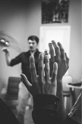 black and white photo of raised hands in focus while a speaker is in the background (image of a talk/lecture)