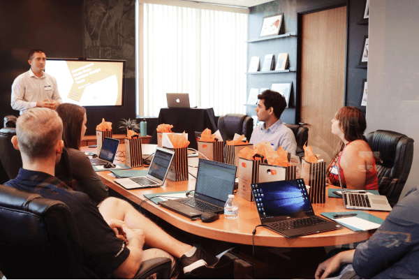 A person presenting in front of the team, laptops out but all are attentive to speaker