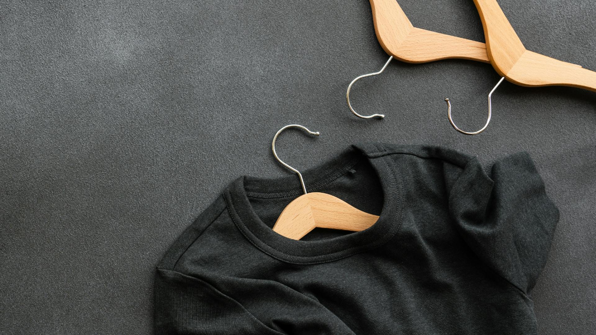 A black t-shirt on a wooden hanger lies on a dark-gray surface. A black t-shirt on a wooden hanger lies on a dark-gray surface.