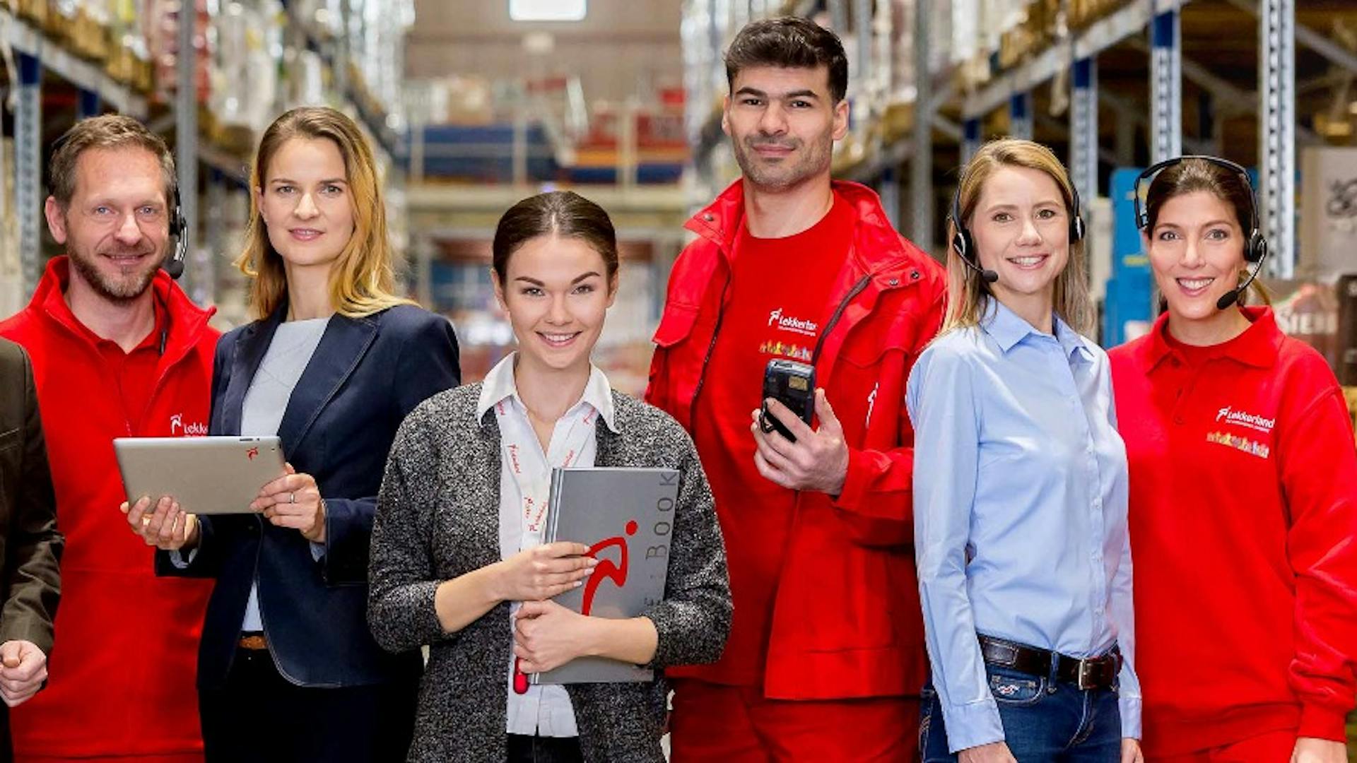 Lekkerland employees stand in a warehouse, all smiling at the camera. Lekkerland employees stand in a warehouse, all smiling at the camera.