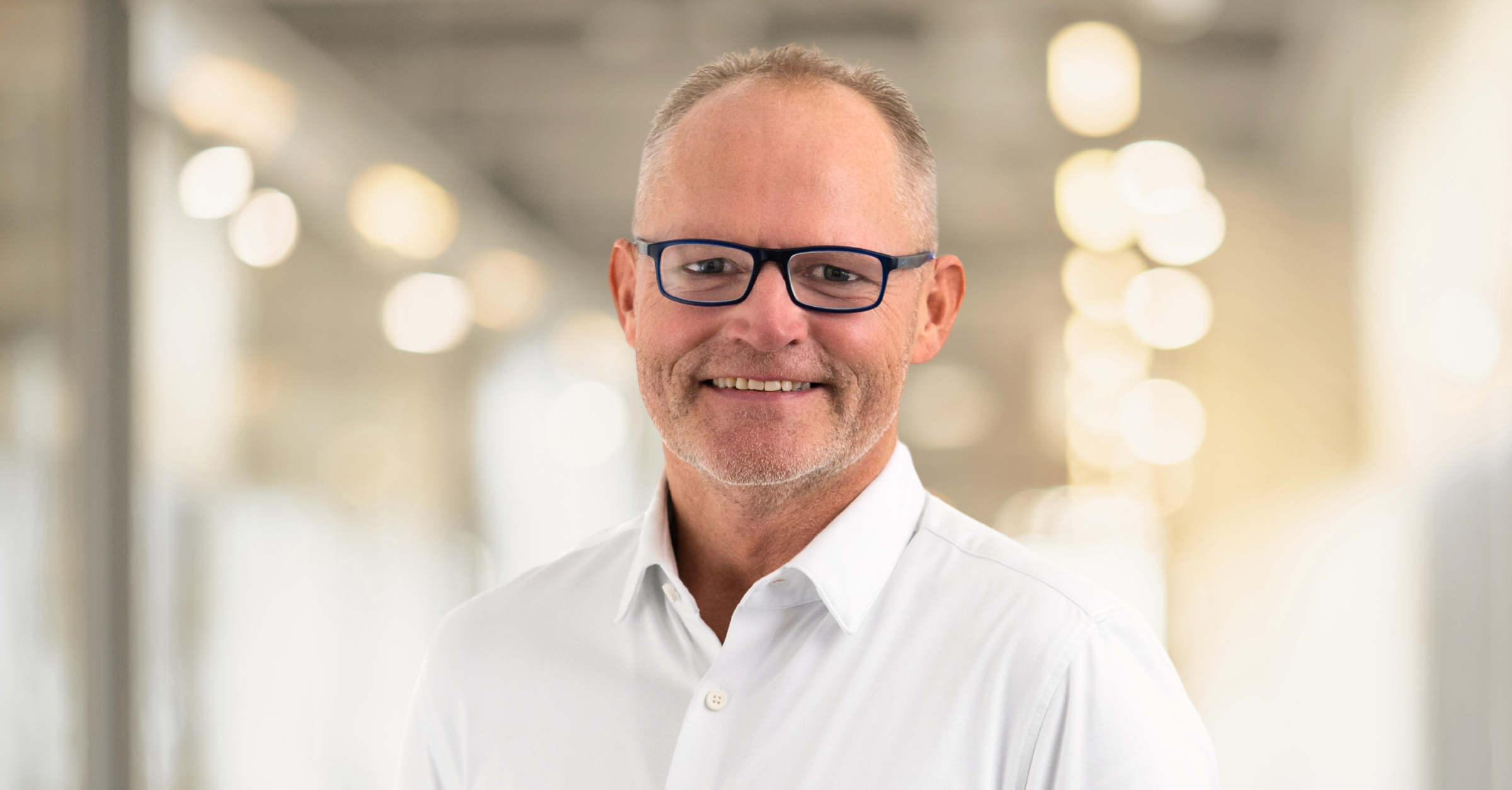Ein Mann mit kurzen blonden Haaren, schwarzer Brille und weißem Hemd steht in einem Raum mit sanfter, verschwommener Beleuchtung im Hintergrund.