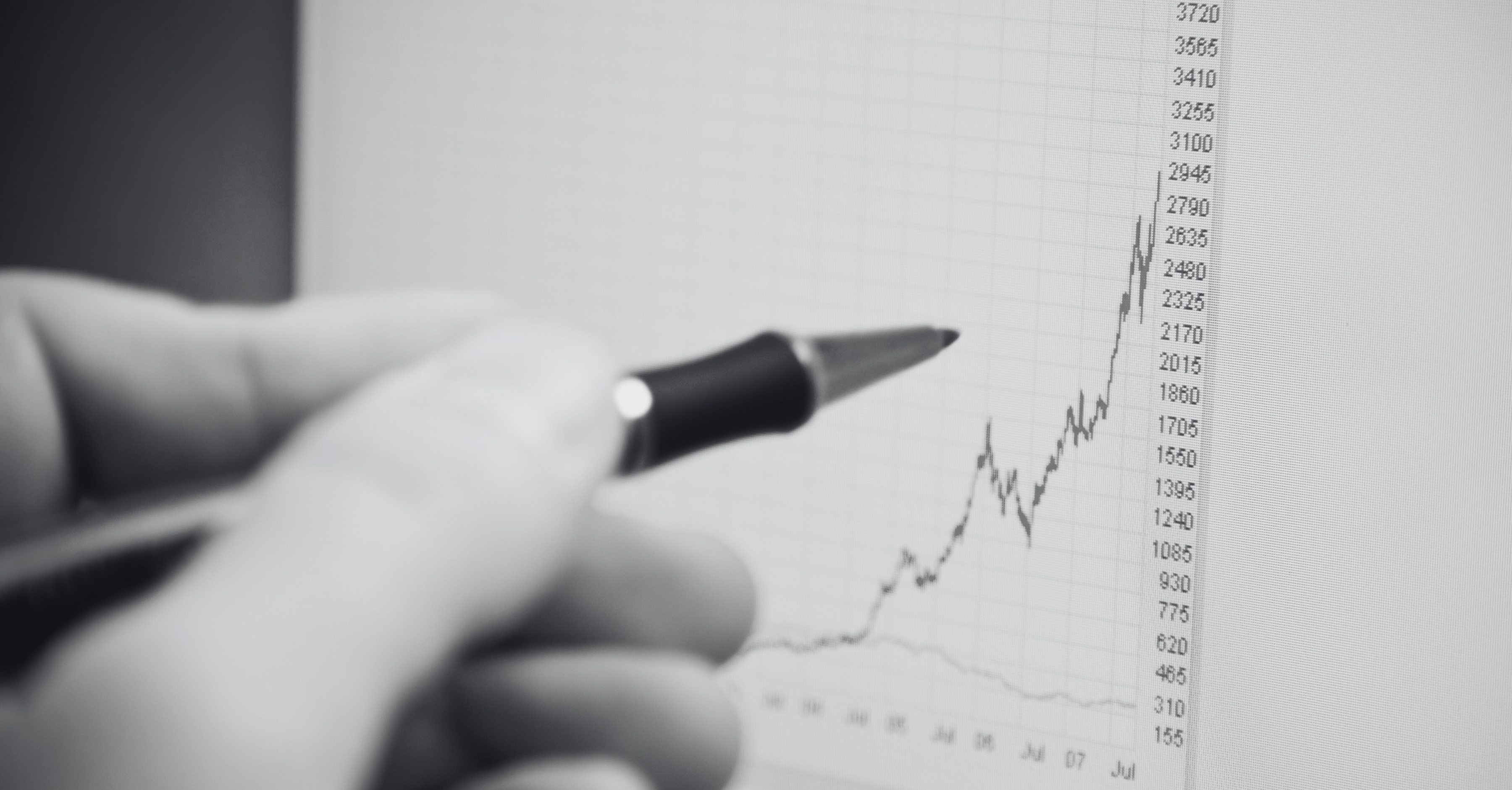 A hand holds a pen and points to a sharply rising line on a financial chart with data points displayed on a screen.