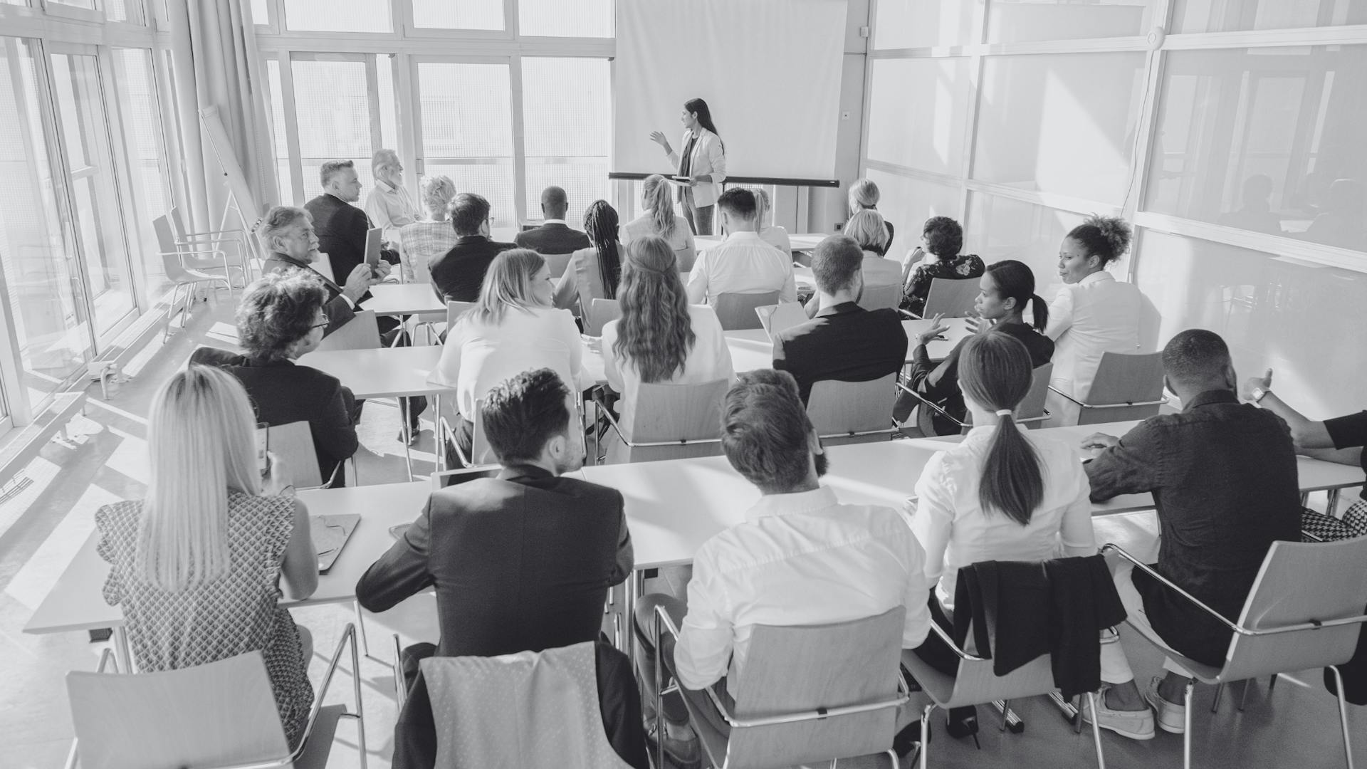 Ein Seminarraum, in dem eine Frau vor sitzenden Personen steht und spricht. Ein Seminarraum, in dem eine Frau vor sitzenden Personen steht und spricht.