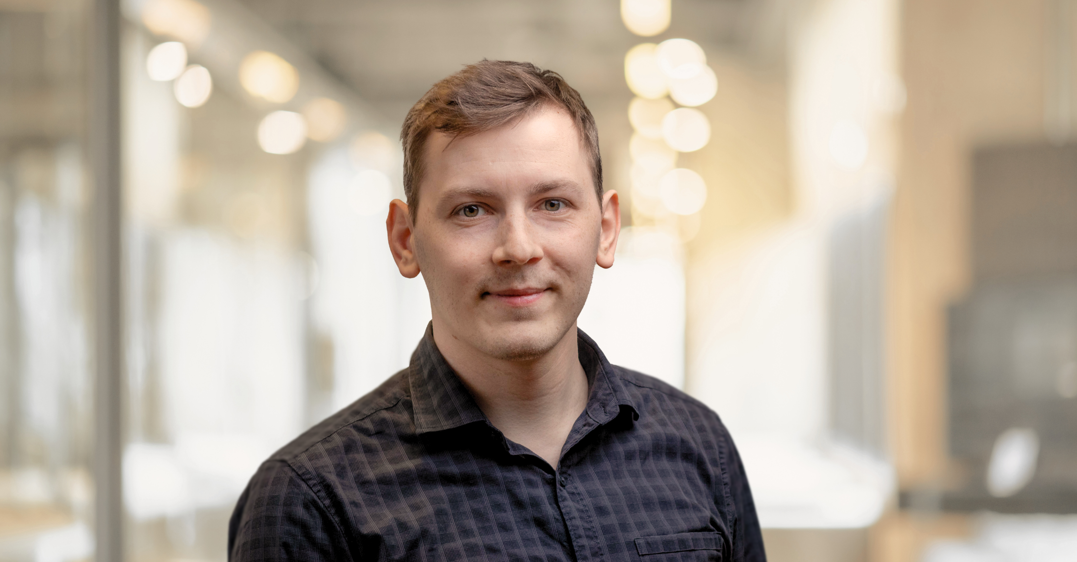 Eine Person mit kurzen Haaren, die ein schwarz kariertes Hemd trägt, steht in einem Innenraum mit sanfter, verschwommener Beleuchtung im Hintergrund.