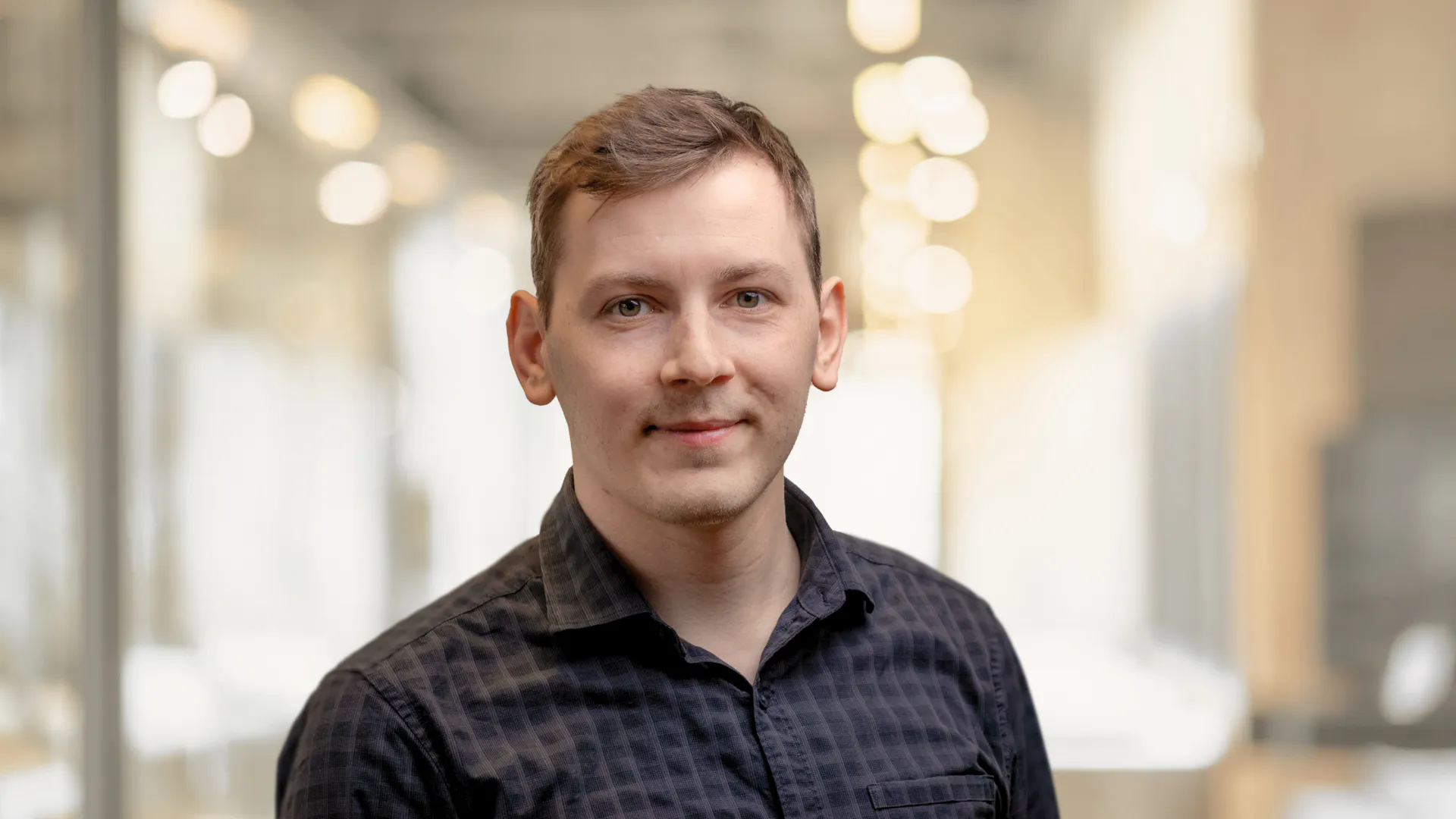 A person with short hair wearing a black checkered shirt stands indoors with soft, blurred lighting in the background. A person with short hair wearing a black checkered shirt stands indoors with soft, blurred lighting in the background.