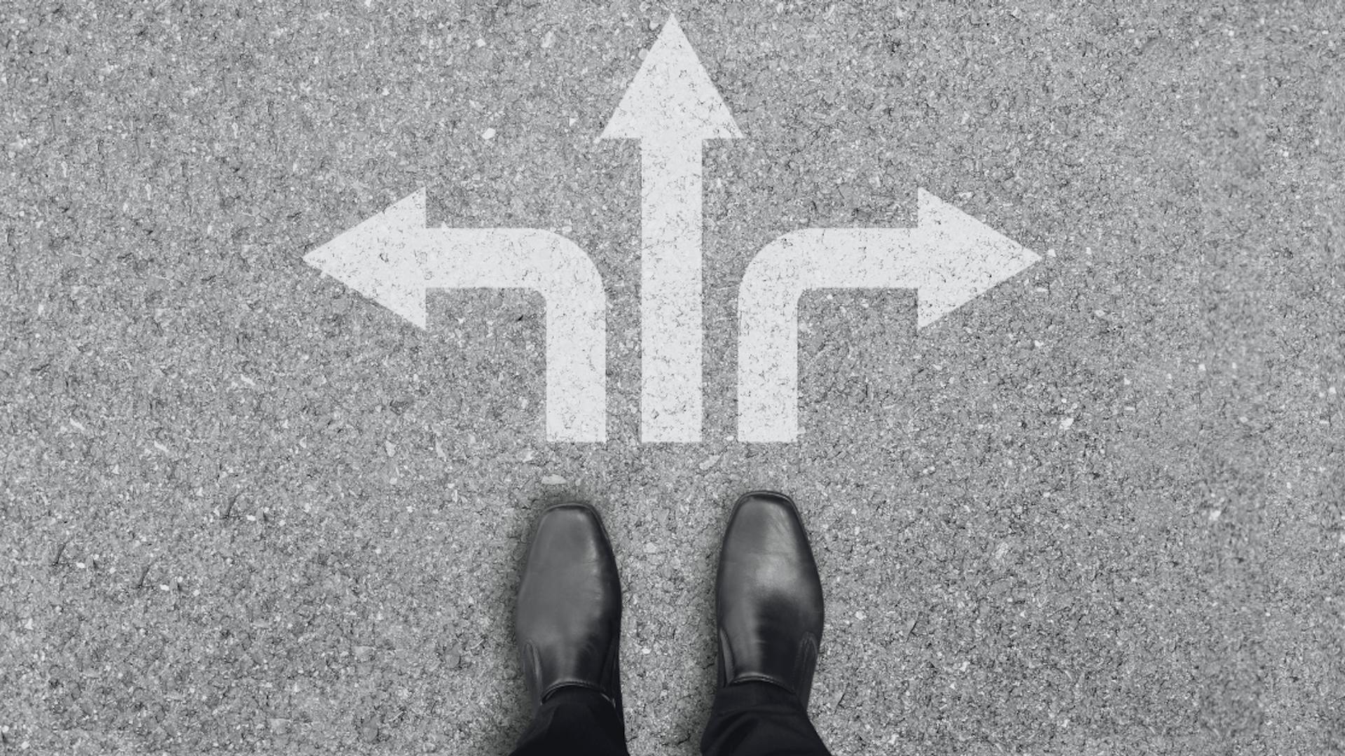 A pair of black shoes stands on a concrete surface with three large white arrows pointing left, straight, and right. A pair of black shoes stands on a concrete surface with three large white arrows pointing left, straight, and right.