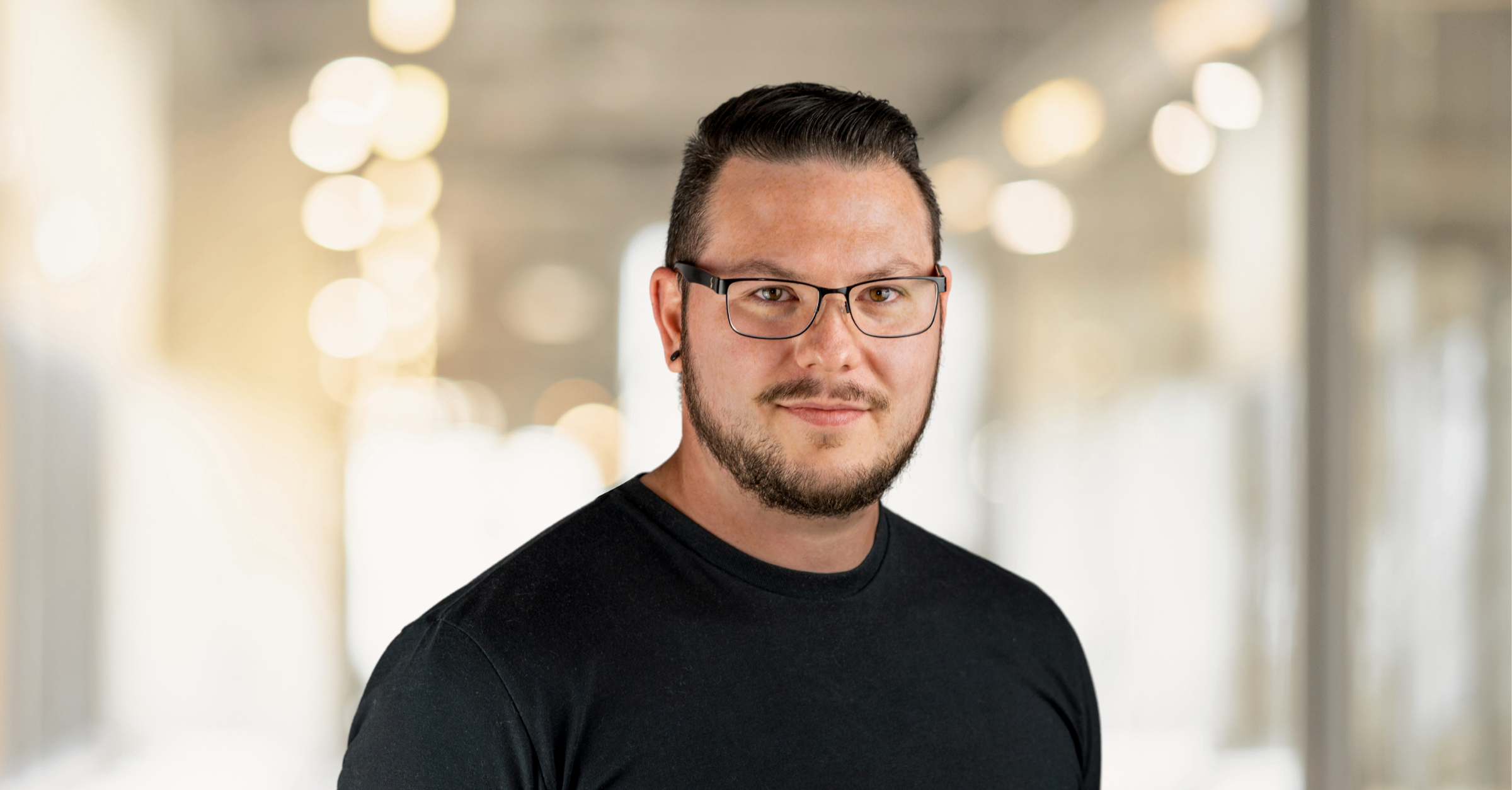 A man with short black hair and a beard wearing a dark shirt stands in a room with soft, blurry lighting in the background.
