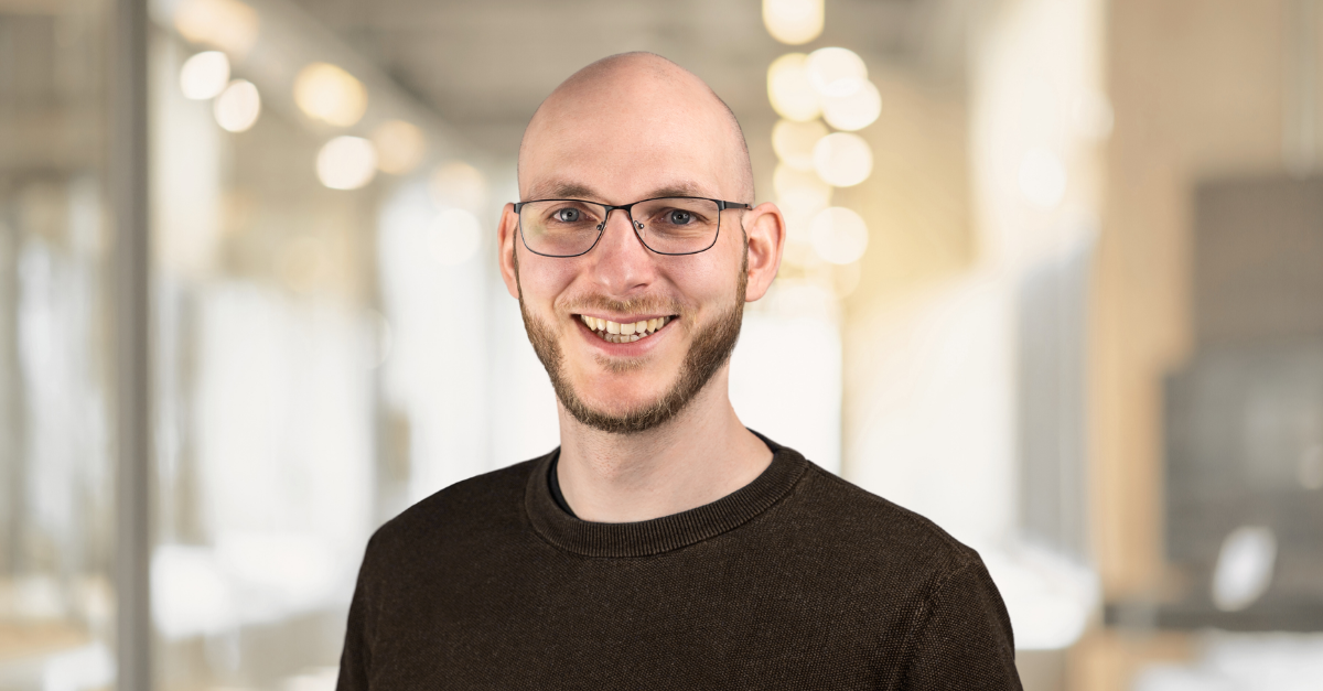 Porträt eines bärtigen Mannes mit Brille und rasiertem Kopf, gekleidet in ein dunkelbraunes Hemd, der in einem Raum mit sanfter, verschwommener Beleuchtung im Hintergrund steht.