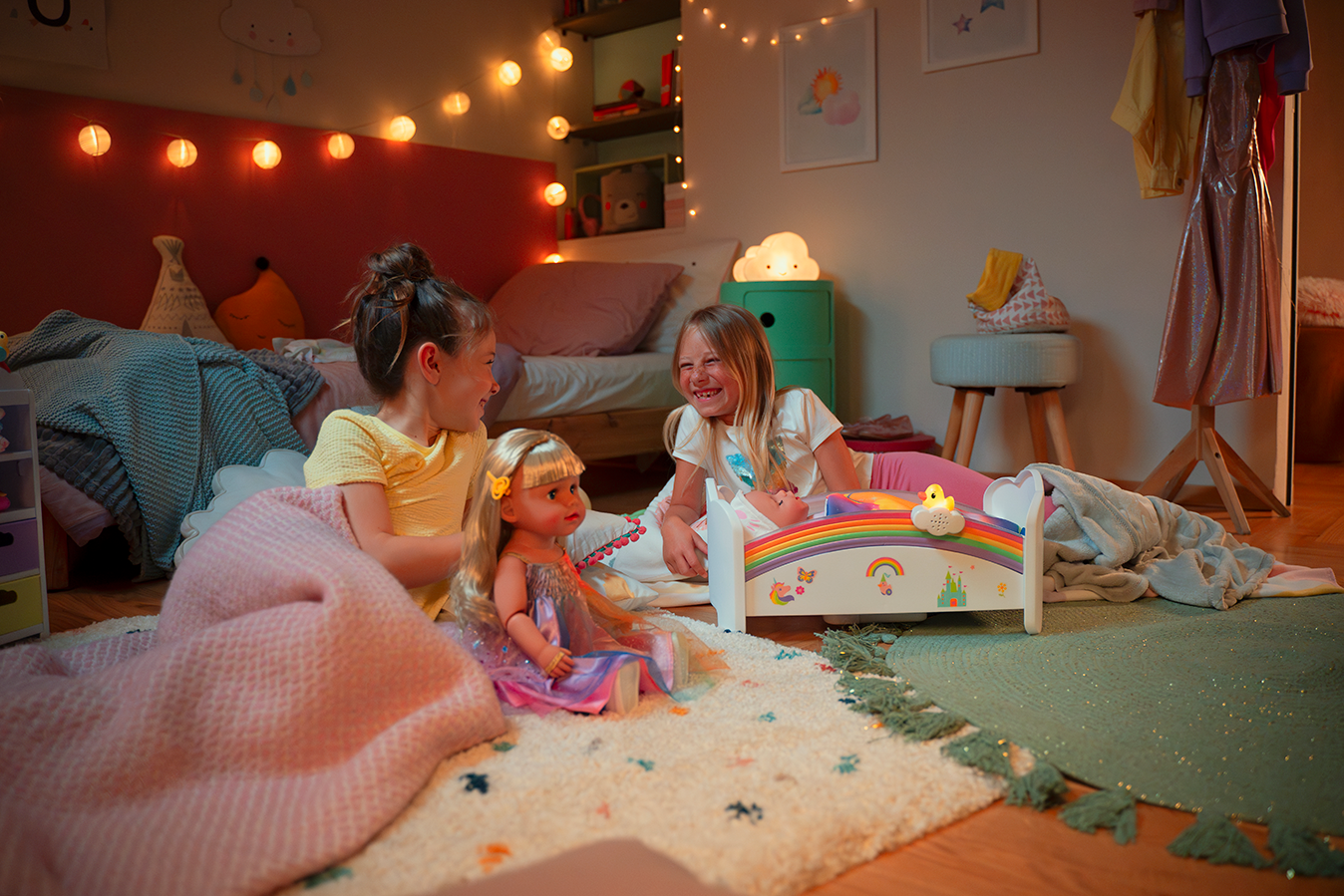 Two girls playing with a BABY born® doll in a cozy bedroom, surrounded by toys and colorful decor.