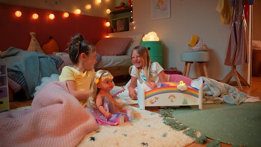Two girls playing with a BABY born® doll in a cozy bedroom, surrounded by toys and colorful decor.