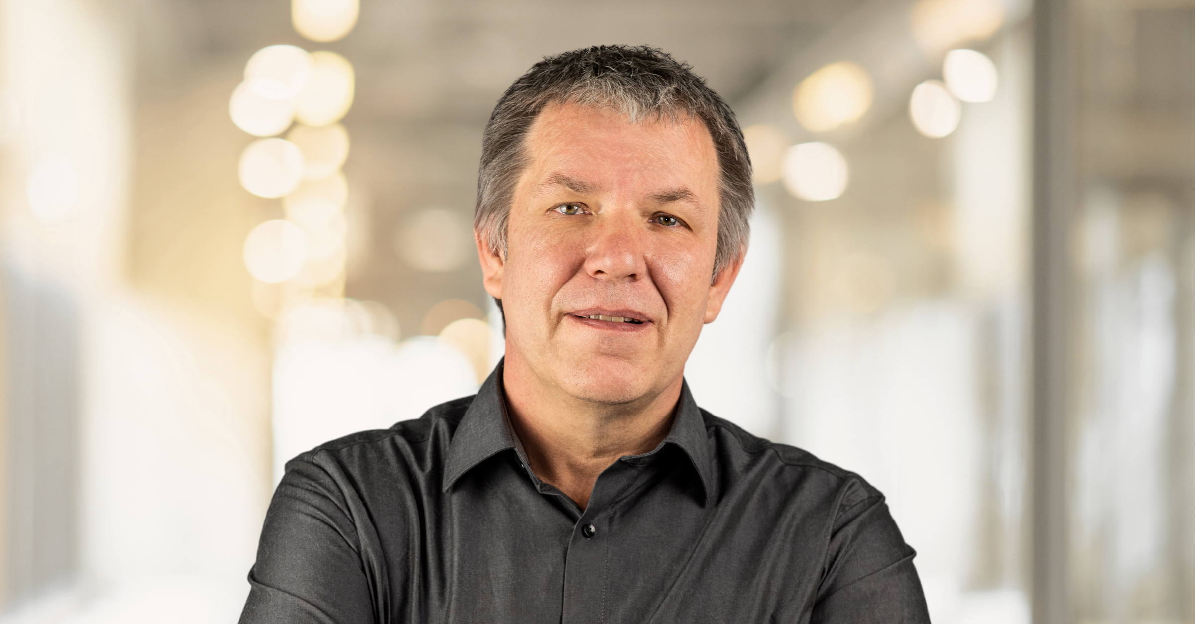 Porträt eines Mannes mit kurzen grauen Haaren, der ein dunkles Hemd mit Knöpfen trägt und mit verschränkten Armen in einem Raum mit sanfter, verschwommener Beleuchtung steht.