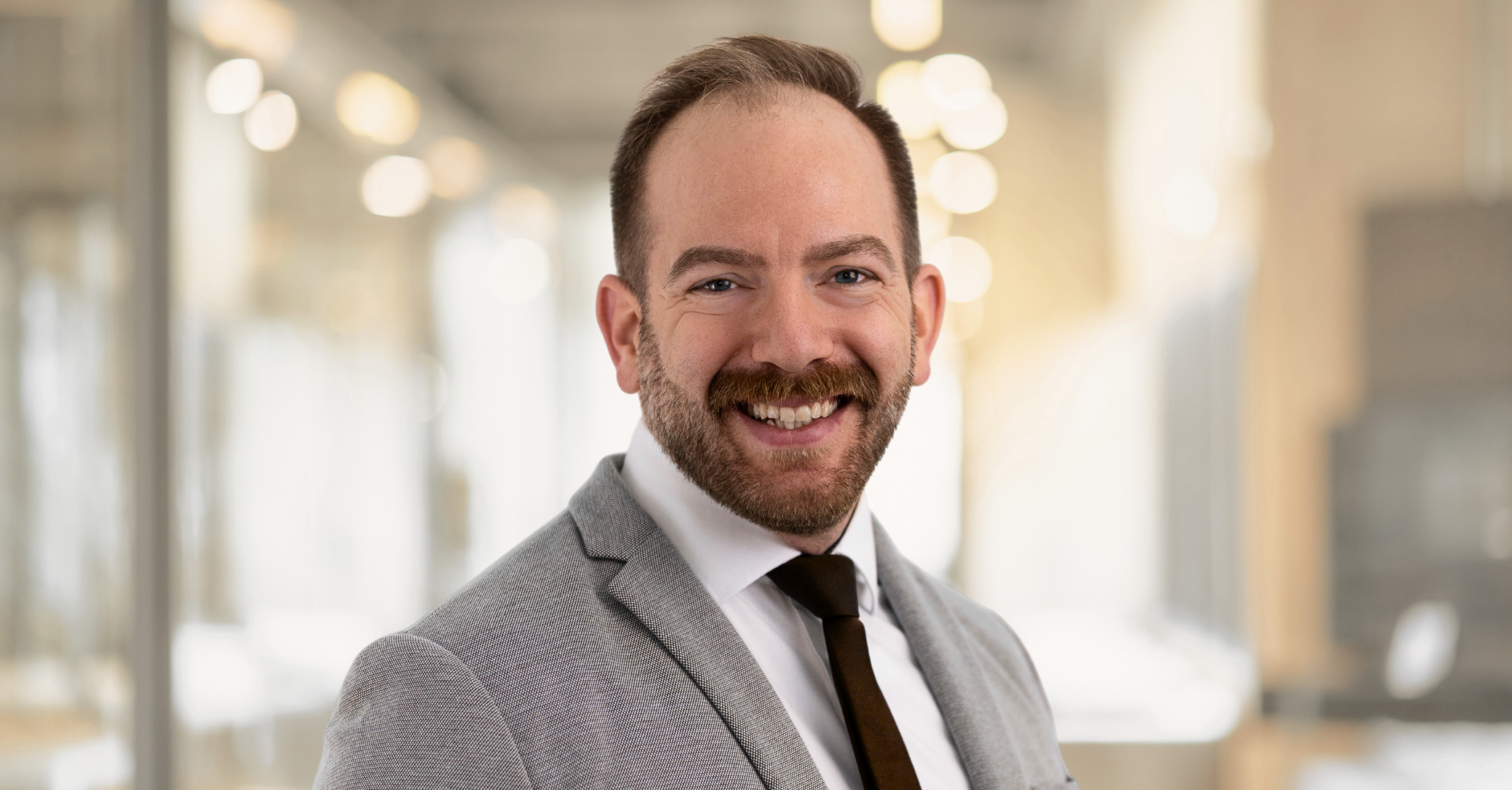 Portrait of a smiling man in a light gray suit and dark tie, standing in a brightly lit modern office with a soft-focus background.