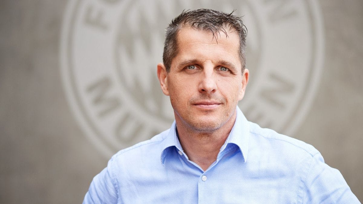 Portrait of Michael Fichtner, CIO at FC Bayern Munich. He has short dark hair and is wearing a light blue button-up shirt. He is standing in front of a soft, blurry grey wall showing a FC Bayern Munich logo.